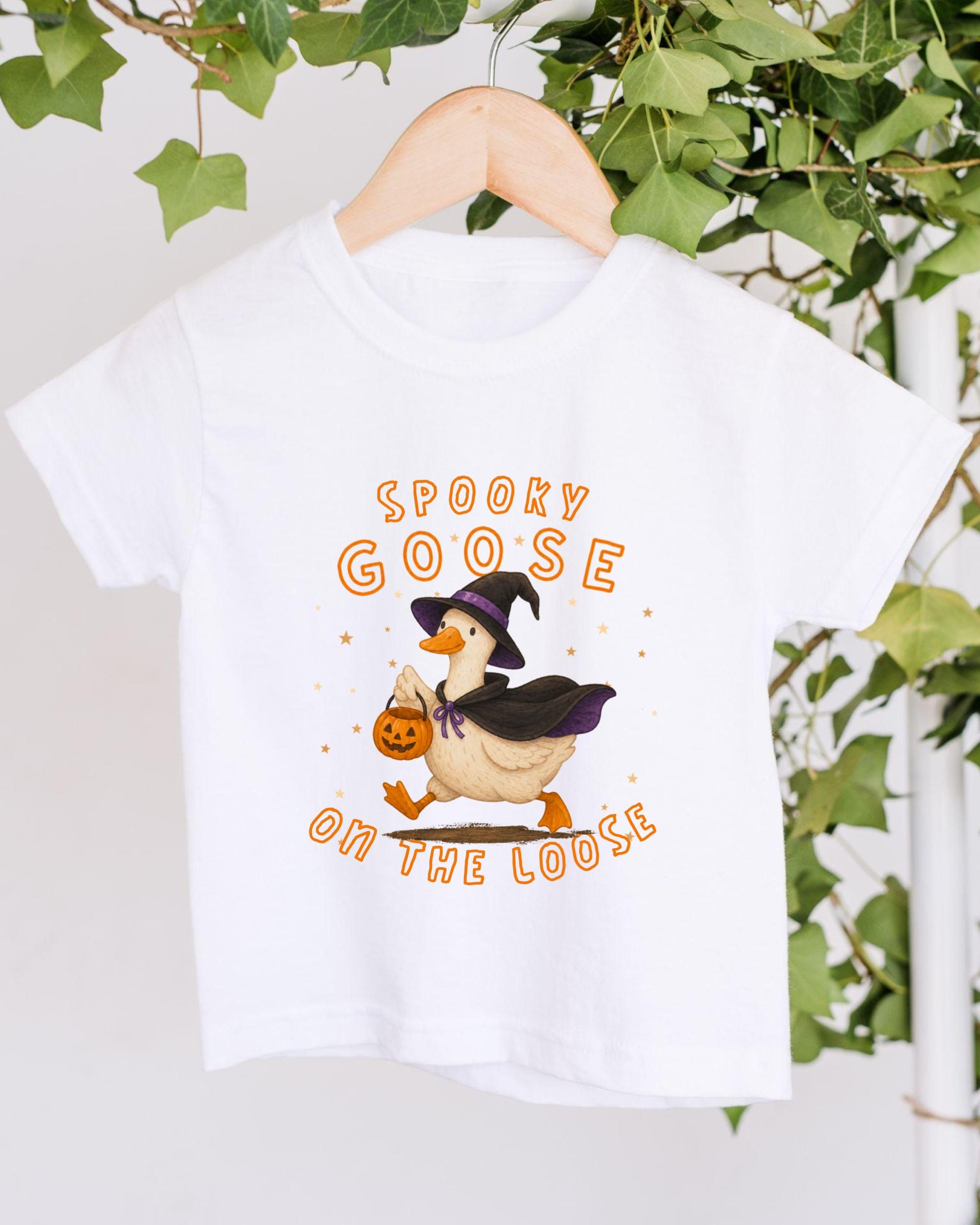 A white t-shirt with a graphic design featuring a cartoon goose dressed as a witch, holding a pumpkin and surrounded by stars. The text on the t-shirt reads "SPOKEY GOOSE ON THE LOOSE".