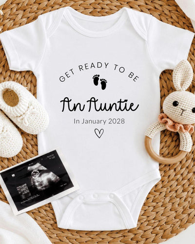 A white baby bodysuit with the text "Get ready to be An Auntie" and "In January 2028" printed on it, along with a pair of white crocheted shoes and a rattle. There is also an image of an Ultrasound showing a baby's feet.