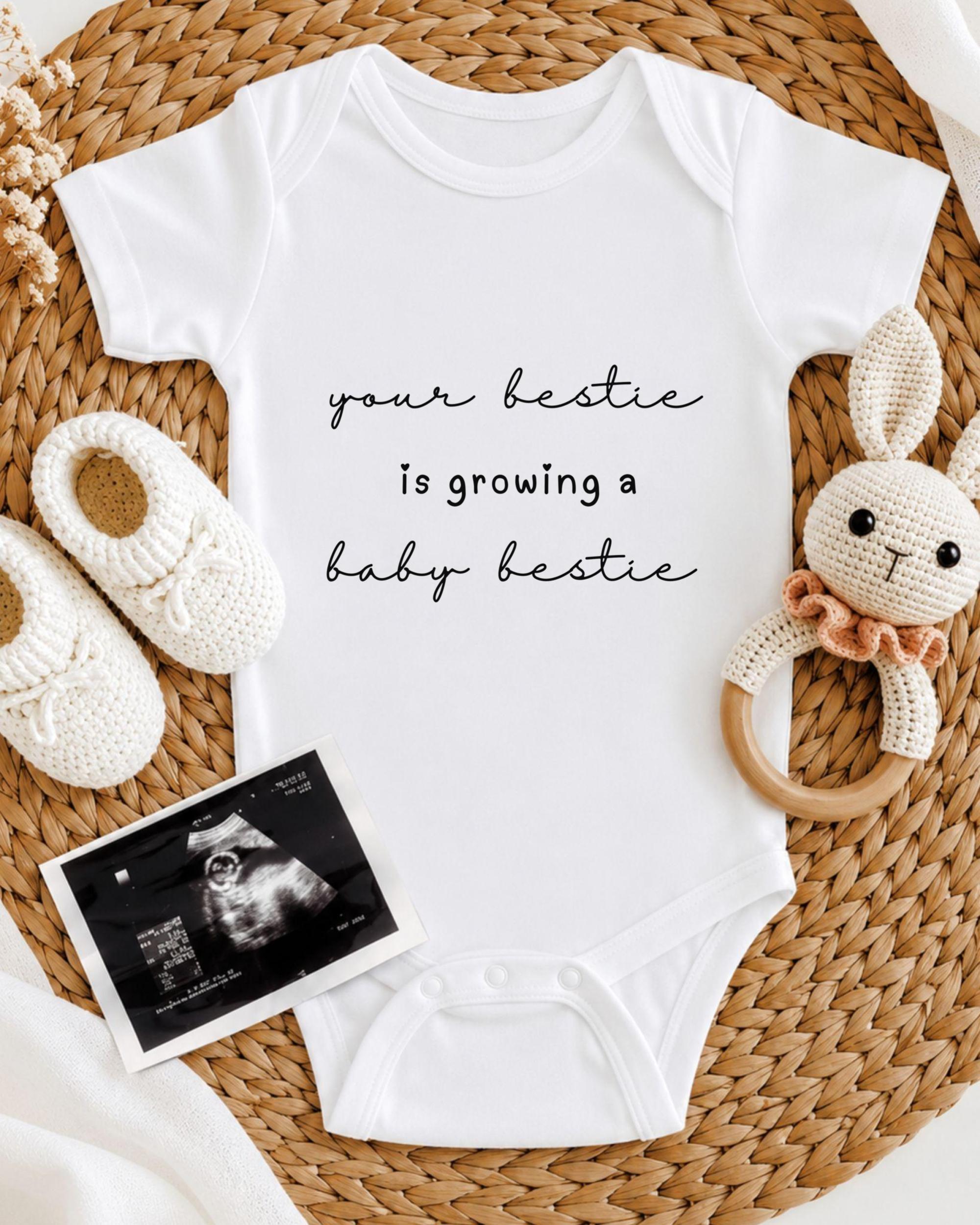 A white onesie with the text "your bestie is growing a baby bestie" printed on it, along with a crocheted bunny toy and a baby shoe. There is also an Ultrasound image of a baby's belly.