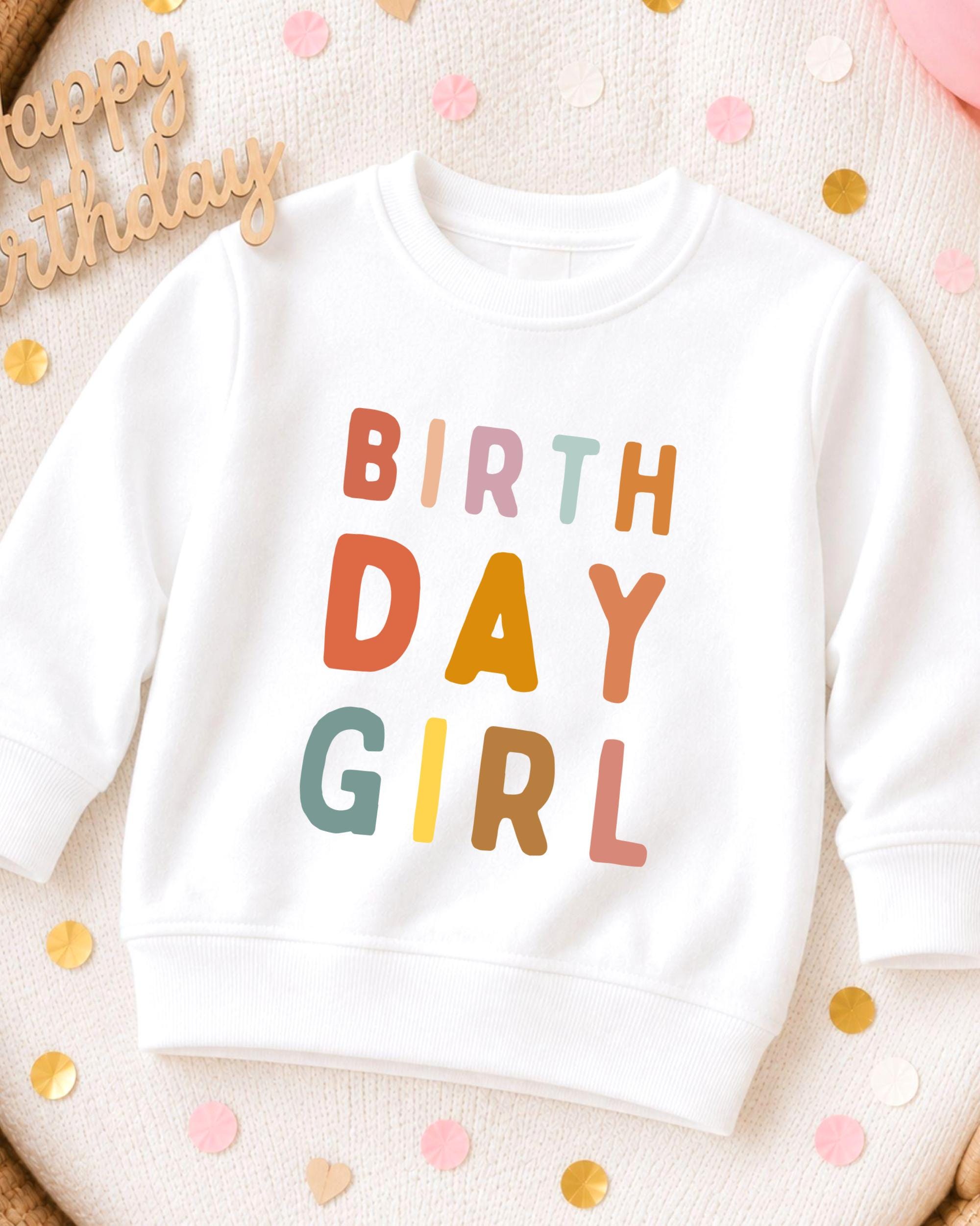 A white sweatshirt with the text "BIRTHDAY GIRL" printed on it, surrounded by colorful confetti.