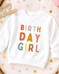 A white sweatshirt with the text "BIRTHDAY GIRL" printed on it, surrounded by colorful confetti.