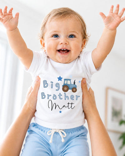 A smiling baby wearing a white shirt with a tractor graphic and the text "Big Brother Matt" is being held up by someone's arms.