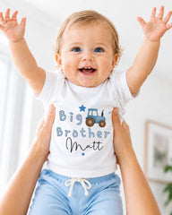 A smiling baby wearing a white shirt with a tractor graphic and the text "Big Brother Matt" is being held up by someone's arms.