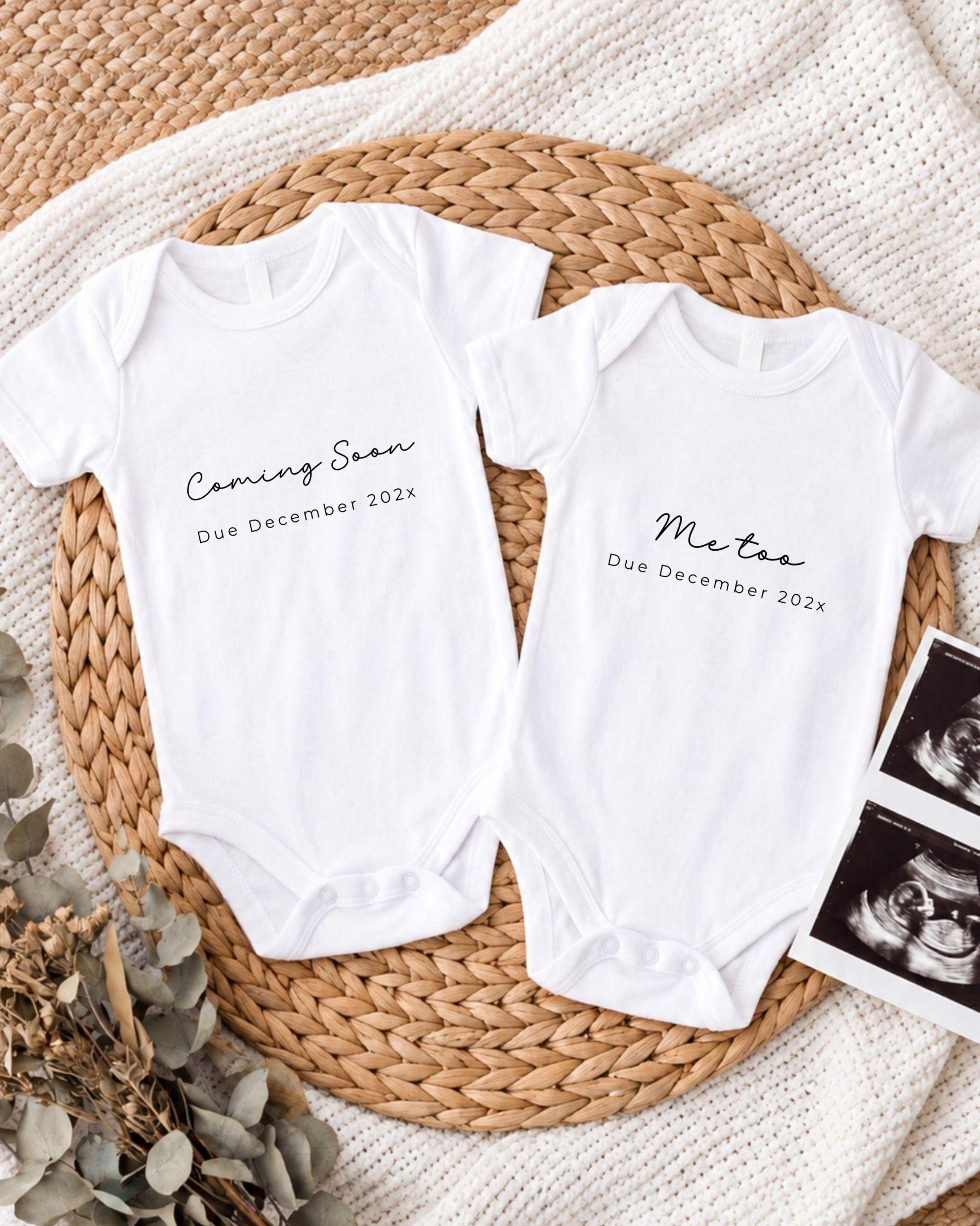 Two white onesies with text announcing the upcoming arrival of a baby, placed on a woven placemat.