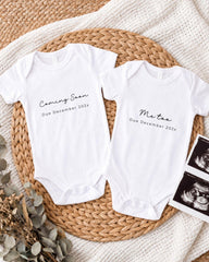 Two white onesies with text announcing the upcoming arrival of a baby, placed on a woven placemat.