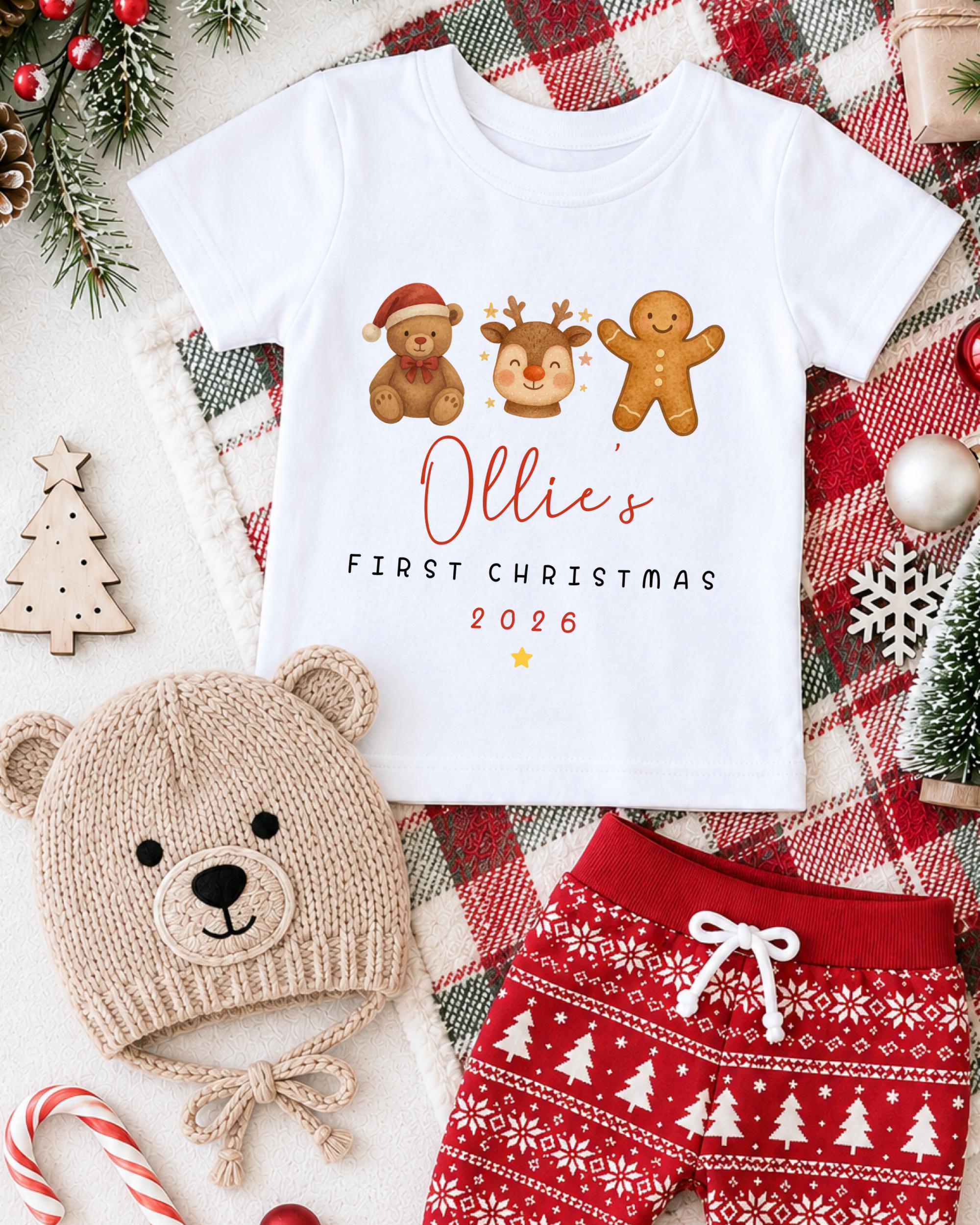 A white t-shirt with a graphic design of three Christmas-themed characters - a teddy bear, a gingerbread man, and a reindeer - along with the text "Ollie's FIRST CHRISTMAS 2006". The t-shirt is displayed on a plaid blanket, and there are other Christmas-themed items such as a knitted hat, a pair of red shorts, and a small Christmas tree in the background.
