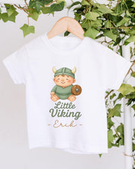 A white t-shirt with a cartoon image of a smiling baby wearing a Viking helmet and holding a shield, along with the text "Little Viking Erik".