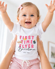 A smiling baby girl wearing a white t-shirt with the text "FIRST TIME FLYER Barcelona" and pink shorts, being held up by someone's arms.