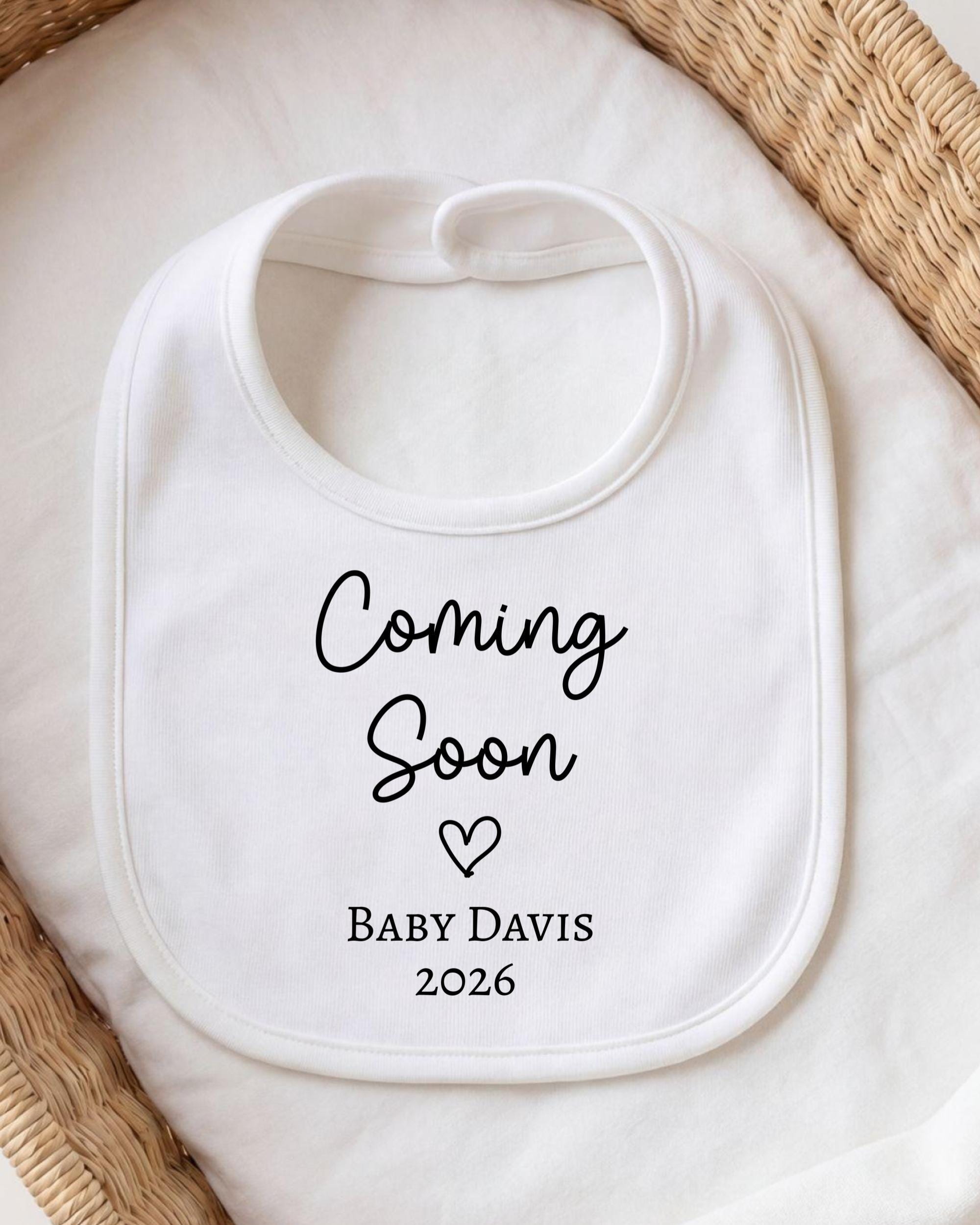 A white bib with the text "Coming Soon" and "Baby Davis 2026" printed on it, placed on a white cloth.