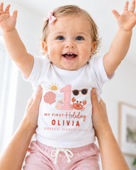 A smiling baby girl wearing a white t-shirt with a personalized design and text, being held up by someone's arms.