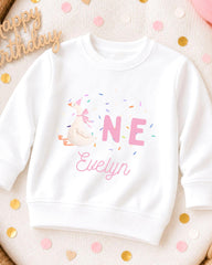 A white sweatshirt with a cartoon goose and the text "ONE" and "Evelyn" printed on it, along with confetti surrounding the sweatshirt.