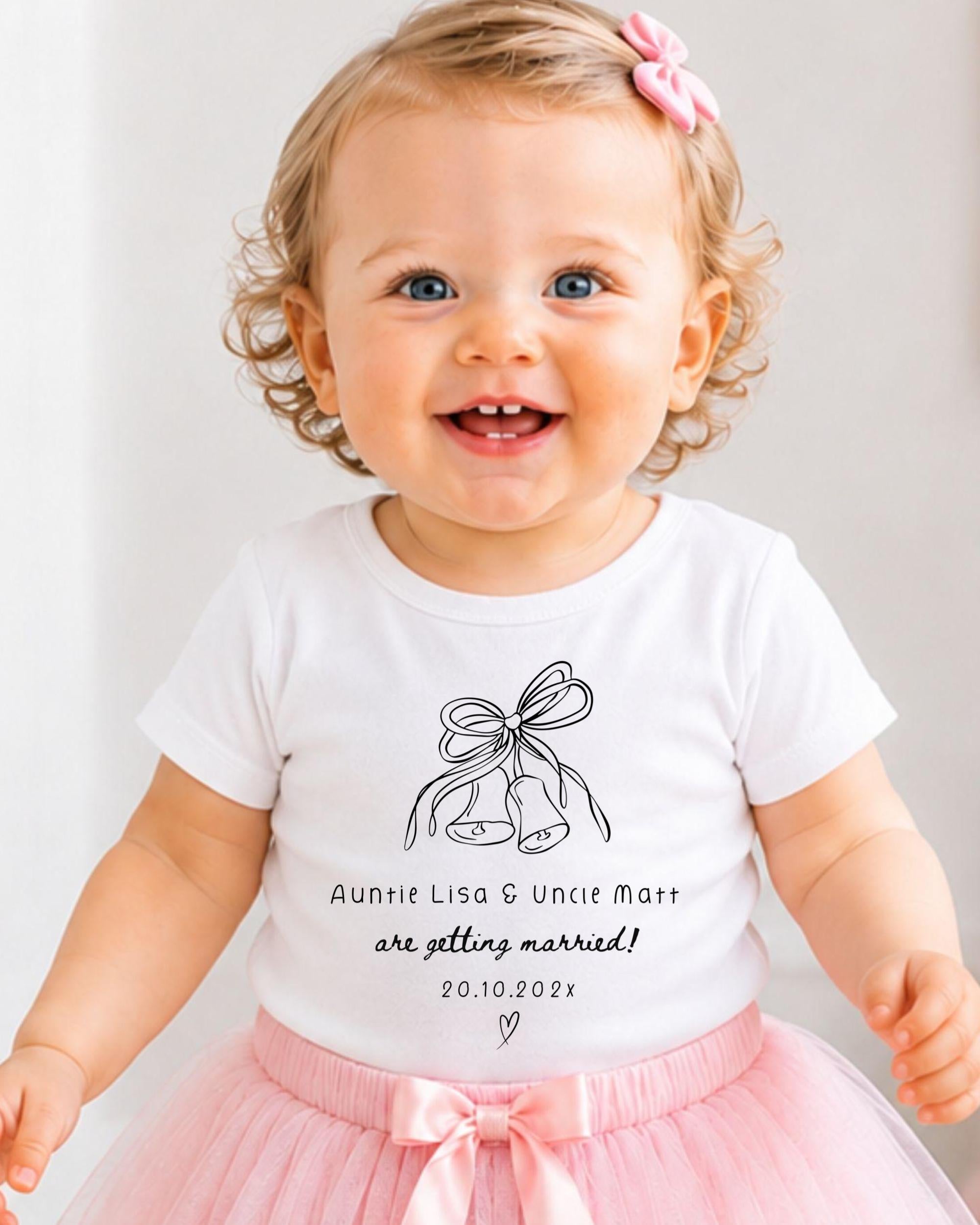 A smiling baby girl wearing a white t-shirt with a graphic and text, standing in front of a white background.