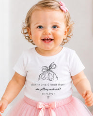 A smiling baby girl wearing a white t-shirt with a graphic and text, standing in front of a white background.