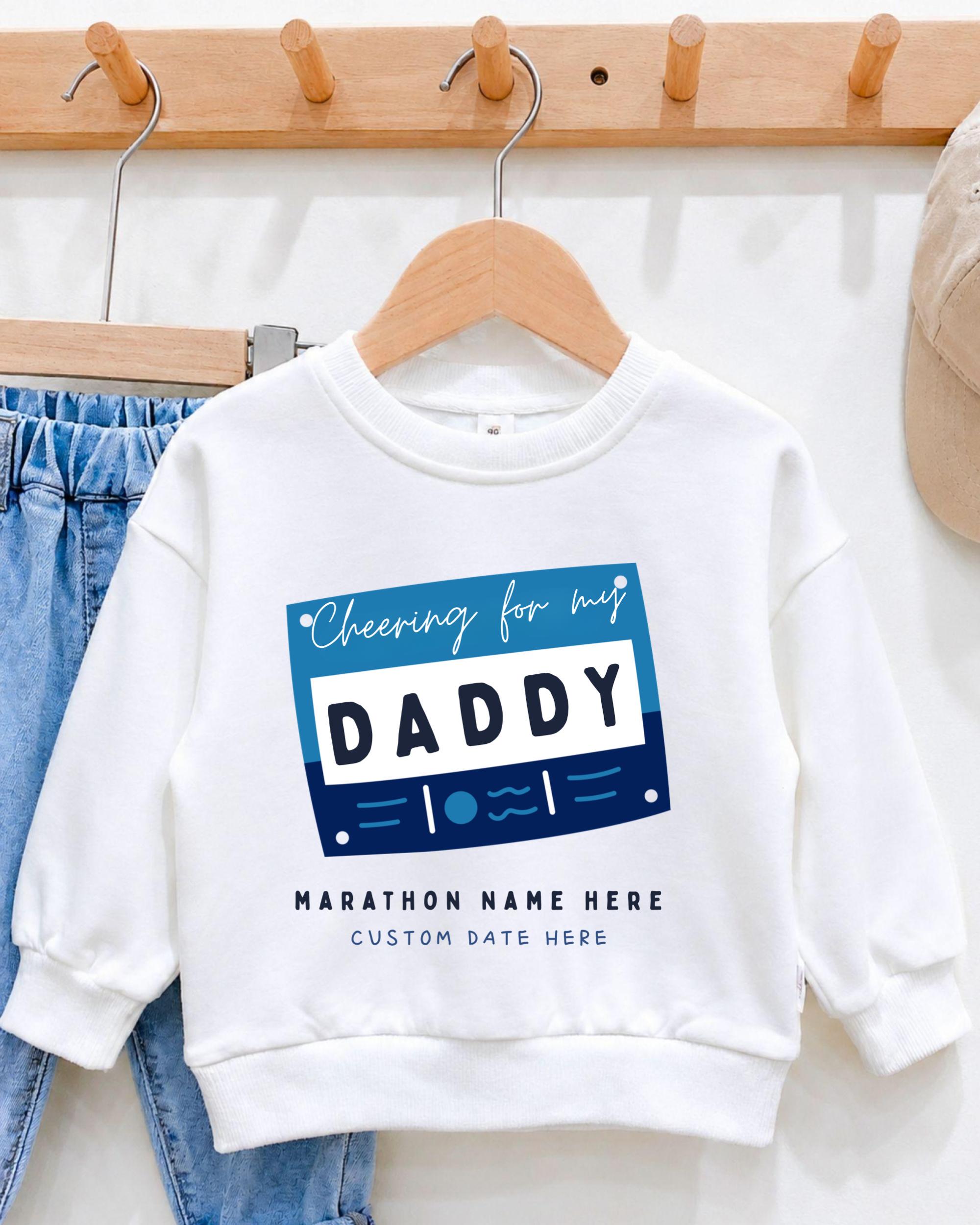 A white sweatshirt with a graphic design that says "Cheering for my DADDY" and "MARCHON NAME HERE" printed on it, hanging on a wooden hanger.