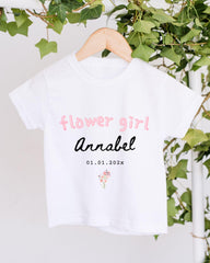 A white t-shirt with pink text and a small floral design hangs on a wooden hanger, surrounded by greenery.