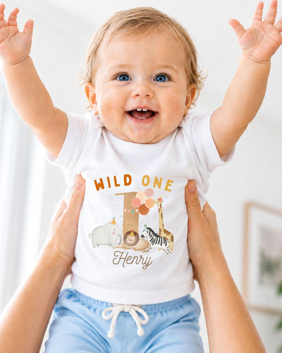 A smiling baby wearing a white t-shirt with a cartoon animal design and the text "Wild One Henry" is being held up by two people.