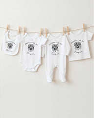 A row of four white baby clothes with the text "PROTECTED BY Cooper" printed on them, hanging on a clothesline.
