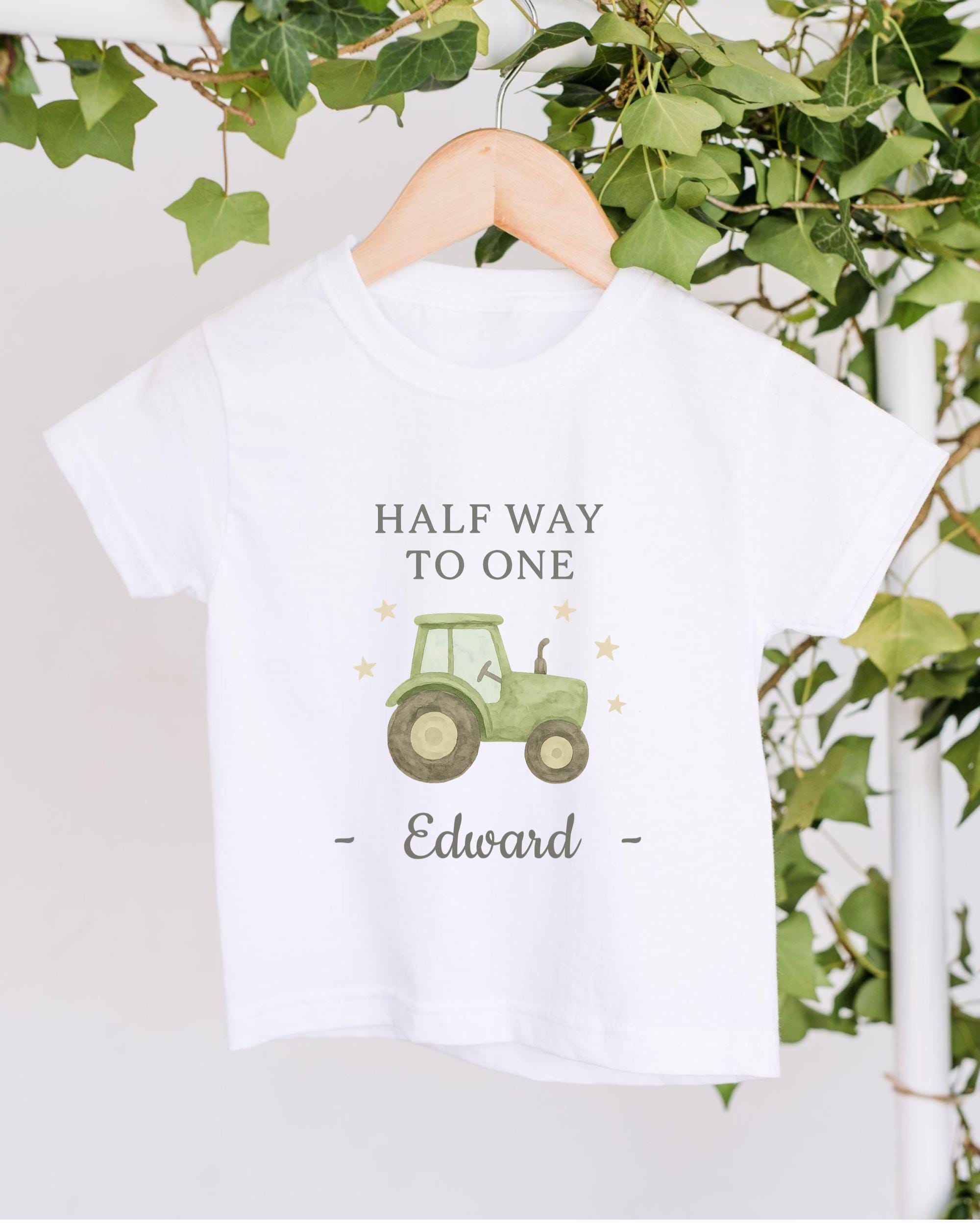 A white t-shirt with a graphic of a tractor and the text "Half Way to One Edward" printed on it, hanging on a wooden hanger.