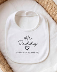 A white bib with the text "Hi Daddy I can't wait to meet you" printed on it, placed on a white cloth.