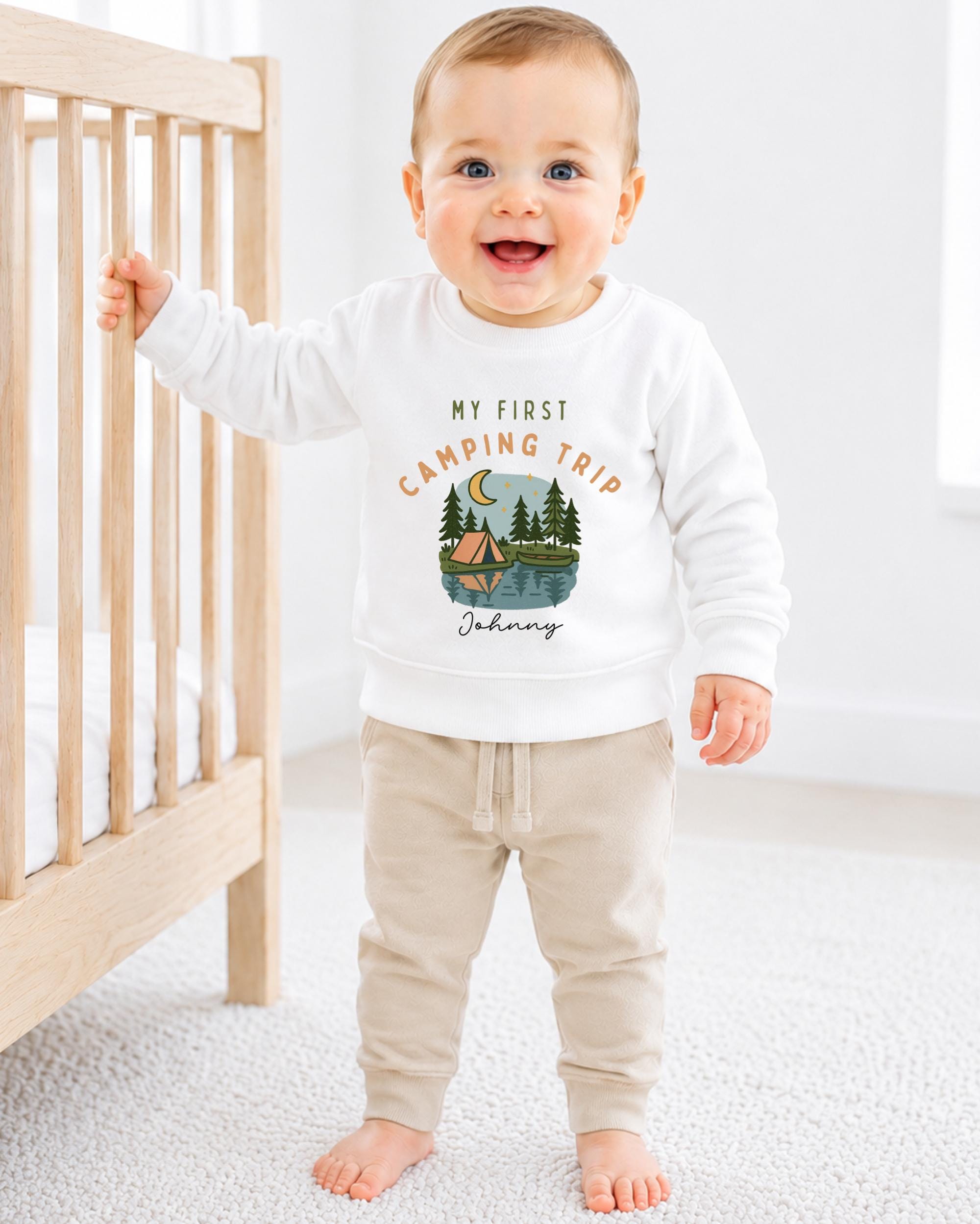 A smiling baby boy wearing a white sweatshirt with a camping trip design and the text "My first camping trip" is standing next to a wooden crib.