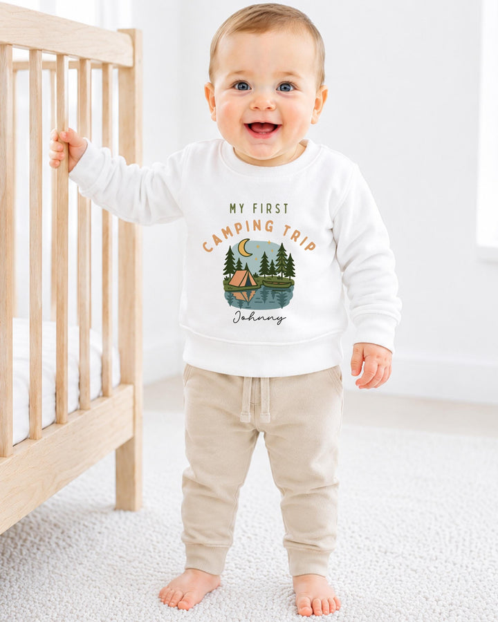A smiling baby boy wearing a white sweatshirt with a camping trip design and the text "My first camping trip" is standing next to a wooden crib.