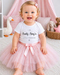A smiling baby girl wearing a white t-shirt with text and a pink tutu sits on a pink carpet, surrounded by stuffed animals and a pink canopy.