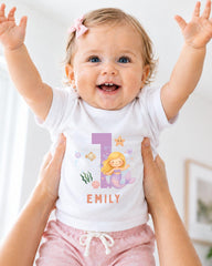 A smiling baby girl wearing a white t-shirt with a mermaid design and the number 1, being held up by someone's arms.