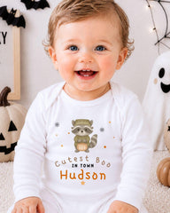 A smiling baby wearing a white onesie with a raccoon graphic and the text "Cute Boo in Town Hudson" printed on it.