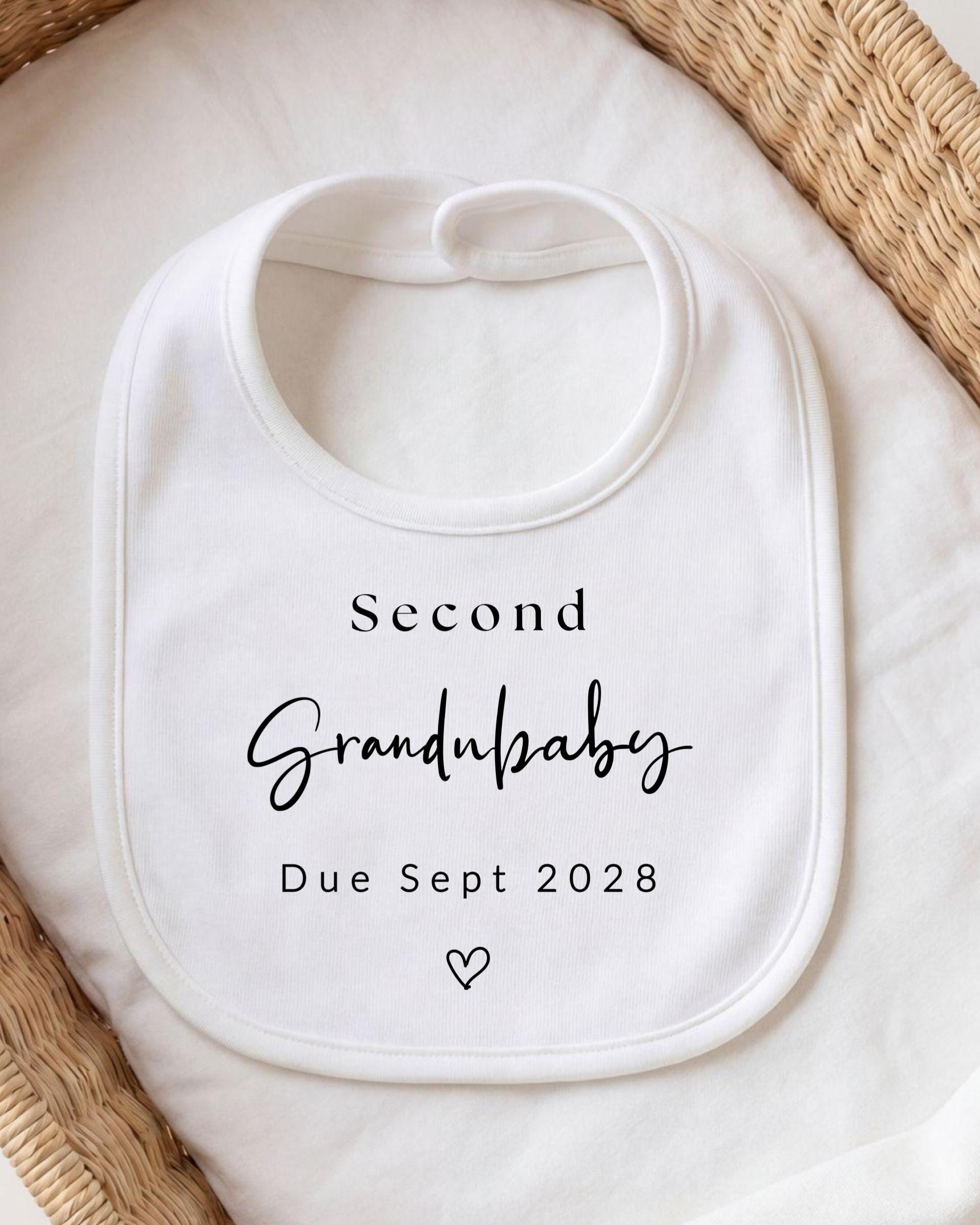 A white bib with the text "Second Grandbaby Due Sept 2028" printed on it, placed on a white cloth.