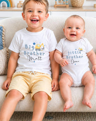 Two young children, a baby and a toddler, sitting on a couch and smiling. They are wearing matching white t-shirts with the text "Big Brother Matt" and "Little Brother Oscar" printed on them.