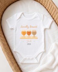 A white onesie with a graphic design of three beer glasses and the text "Locally Brewed Jack 2026" printed on it, placed on a white cloth.