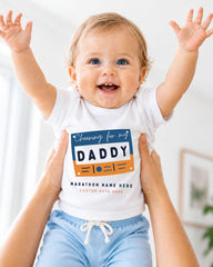 A smiling baby wearing a white shirt with a graphic design and text, being held up by someone's arms.