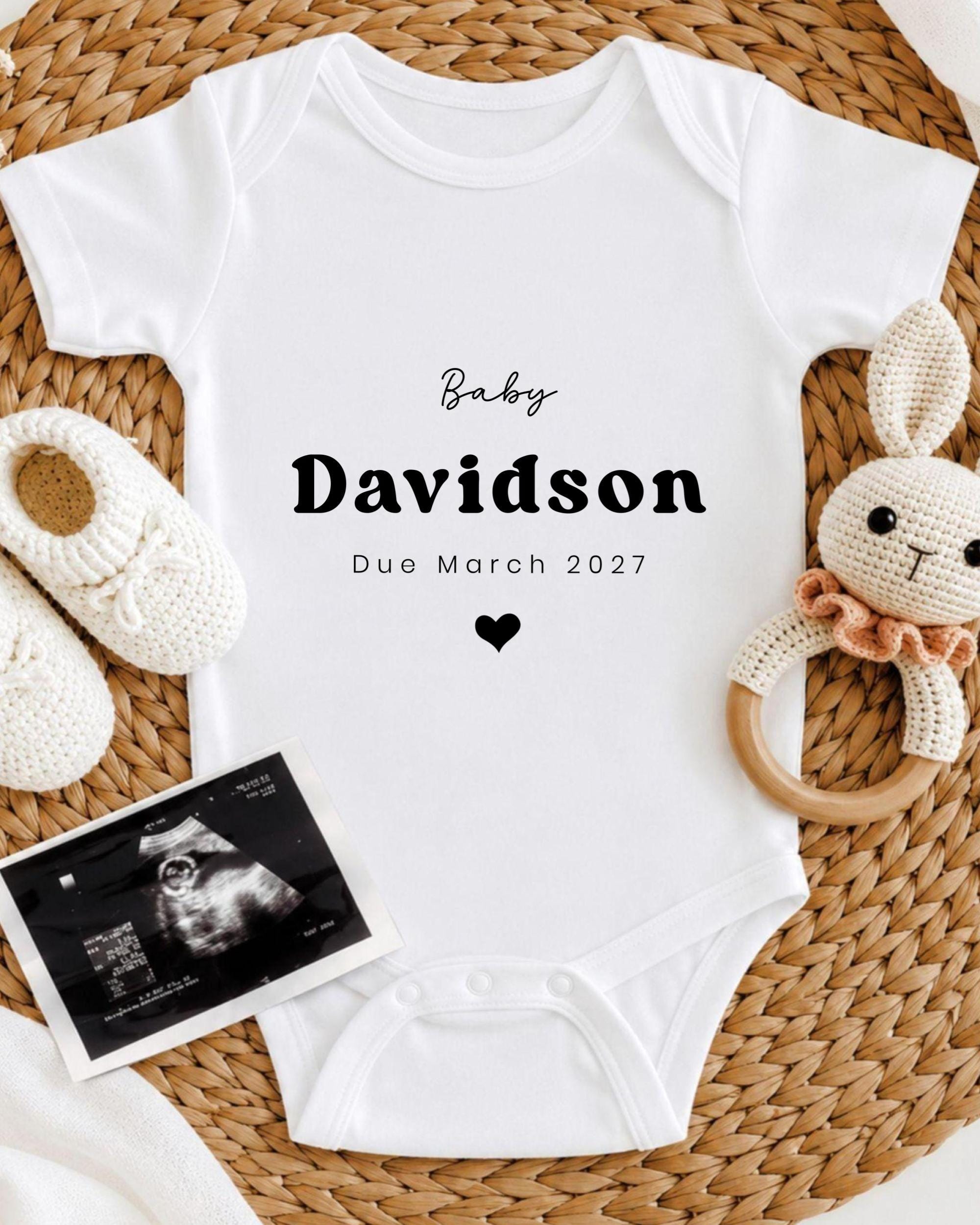 A white baby onesie with the text "Baby Davidson Due March 2027" printed on it, along with a crocheted bunny toy and a baby shoe.