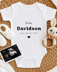 A white baby onesie with the text "Baby Davidson Due March 2027" printed on it, along with a crocheted bunny toy and a baby shoe.