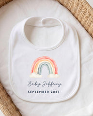 A white bib with a rainbow graphic and the text "Baby Jeffrey September 2027" printed on it.
