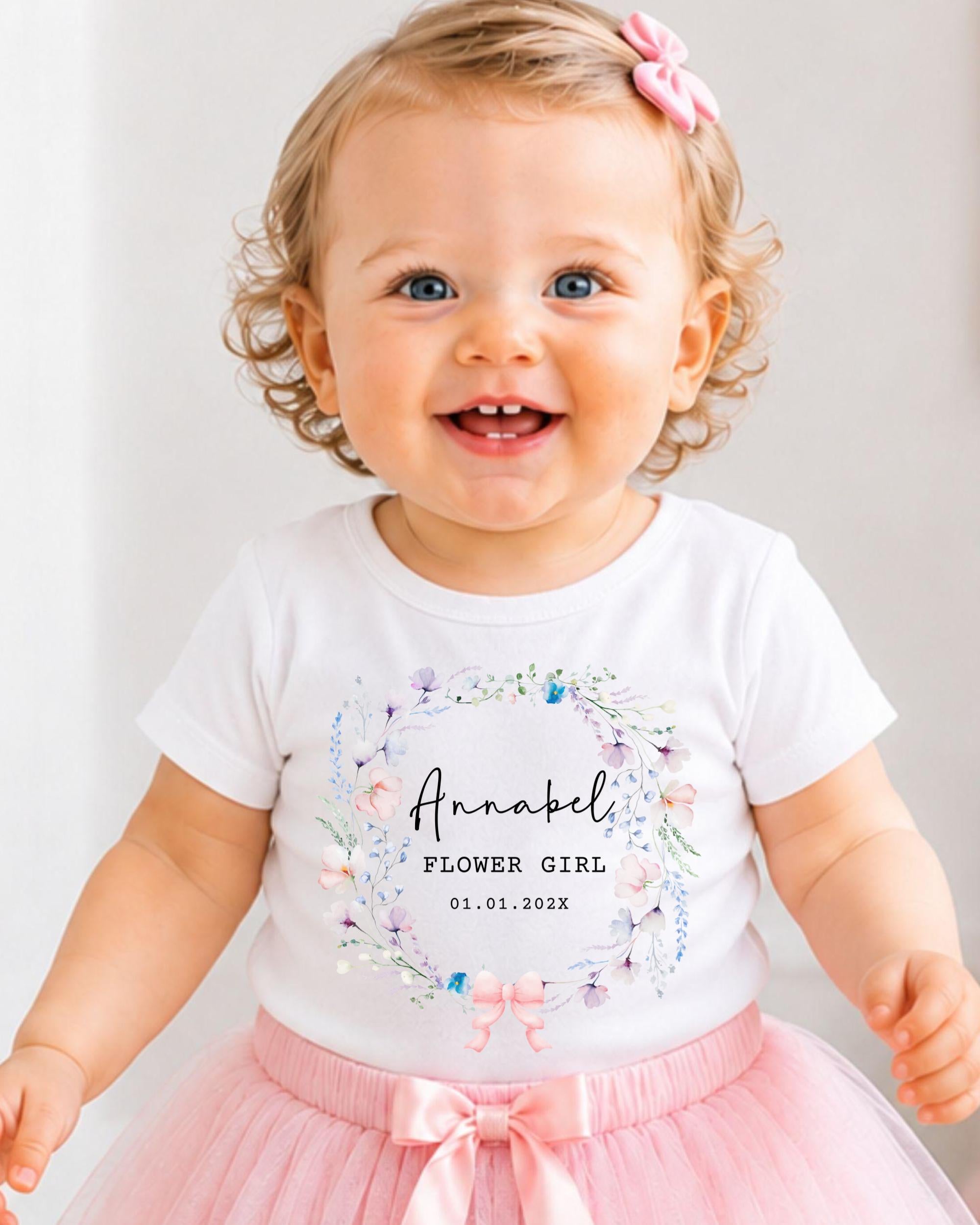 A smiling baby girl wearing a white t-shirt with a floral design and a pink bow in her hair.
