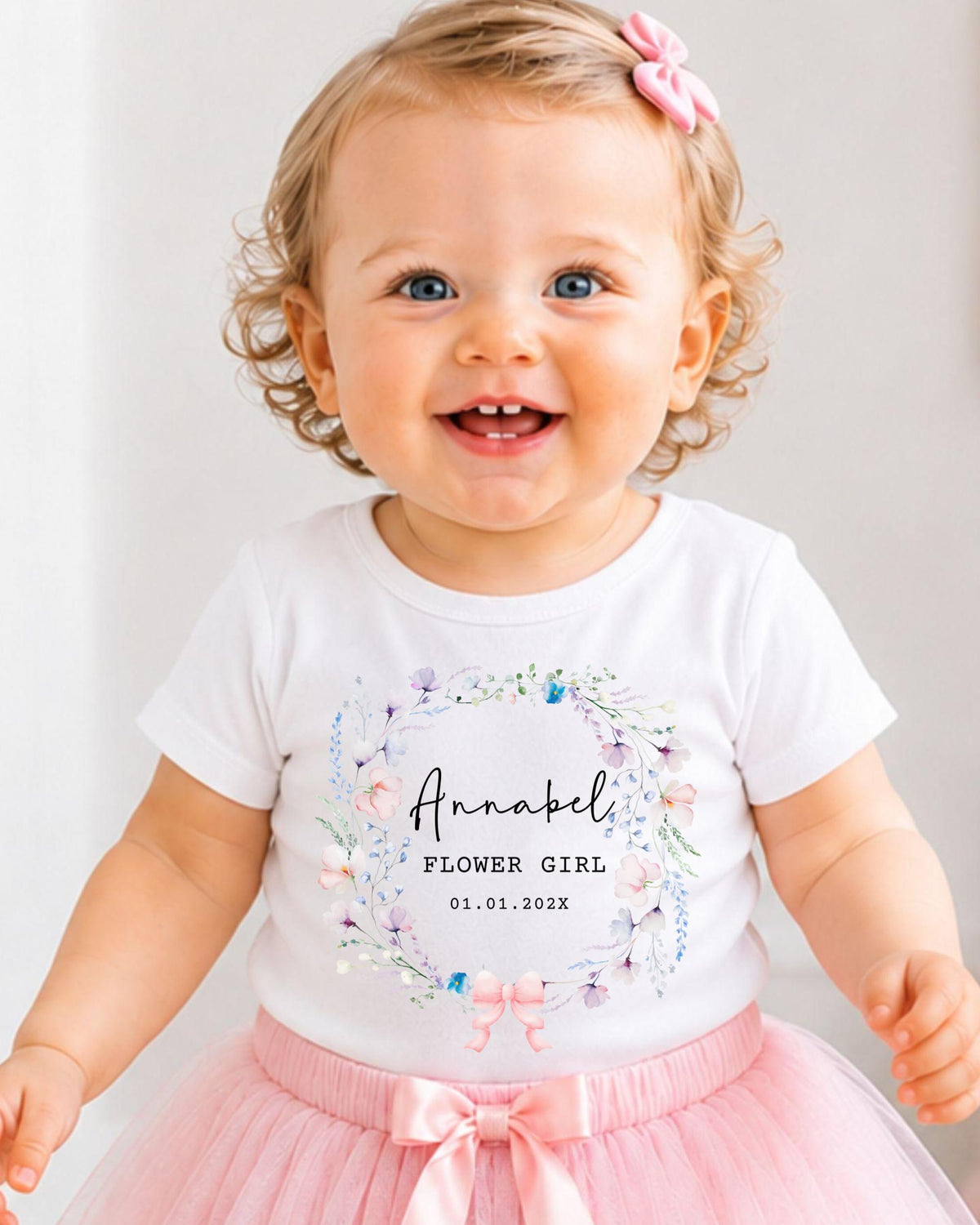A smiling baby girl wearing a white t-shirt with a floral design and a pink bow in her hair.