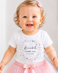 A smiling baby girl wearing a white t-shirt with a floral design and a pink bow in her hair.