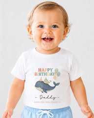 A smiling baby boy wearing a white t-shirt with a graphic of a whale and the text "Happy Birthday Daddy" printed on it.