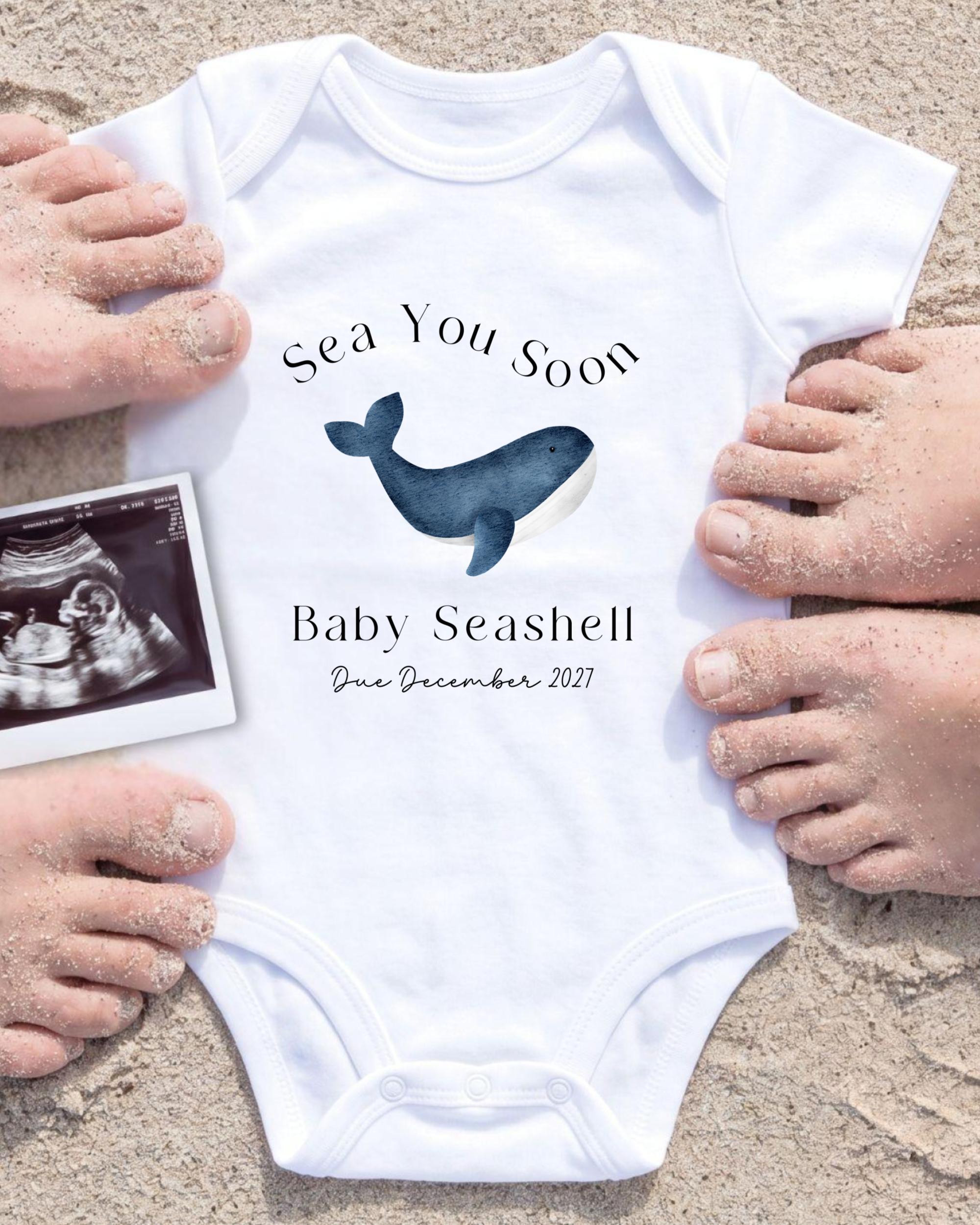 A white baby onesie with a blue whale graphic and text, surrounded by sand and a black and white photograph of a baby's feet.