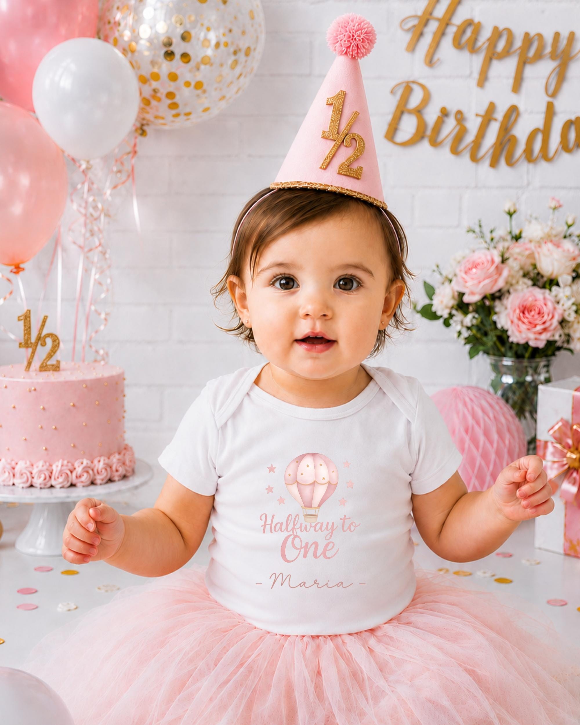 A young girl wearing a white t-shirt with a balloon design and a pink party hat sits in front of a birthday cake and balloons, with a "Happy Birthday" sign in the background.