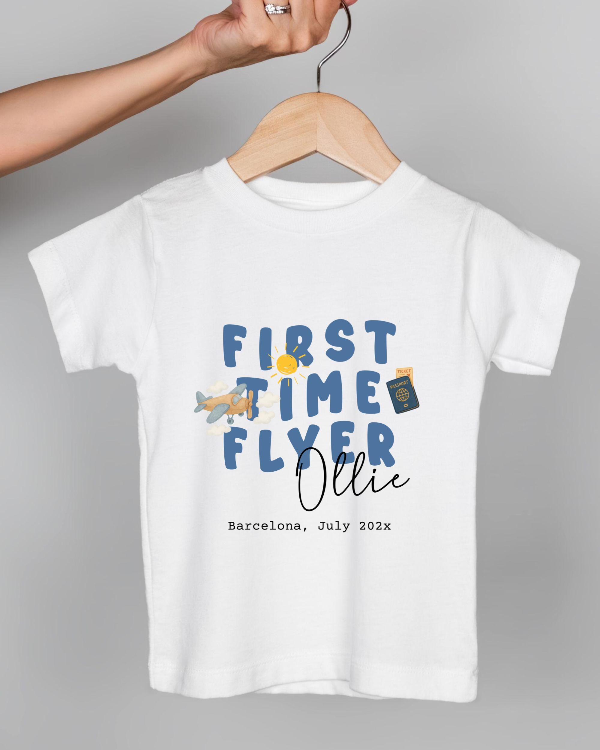 A white t-shirt with a graphic design featuring the text "FIRST TIME FLYER OLLIE" and a small airplane. The text is in blue and orange colors, and the shirt is being held by a person's hand.