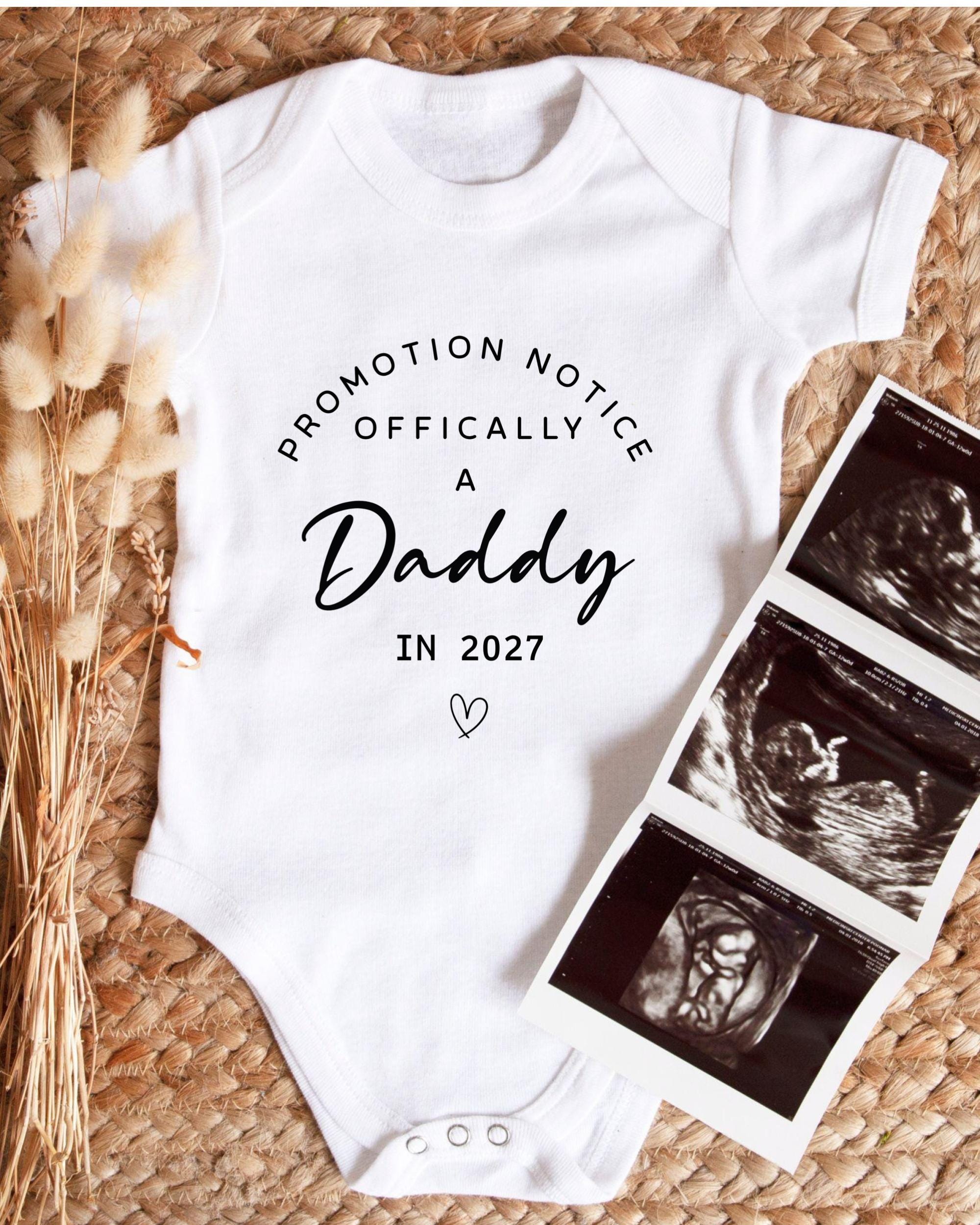 A white baby onesie with text that says "Promotion Notice Officially A Daddy In 2027" and a heart symbol, along with a photograph of an ultrasound image of a baby.
