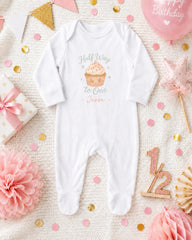 A white onesie with a cupcake design, surrounded by confetti, a pink star, and other baby-related items, all arranged on a white background.