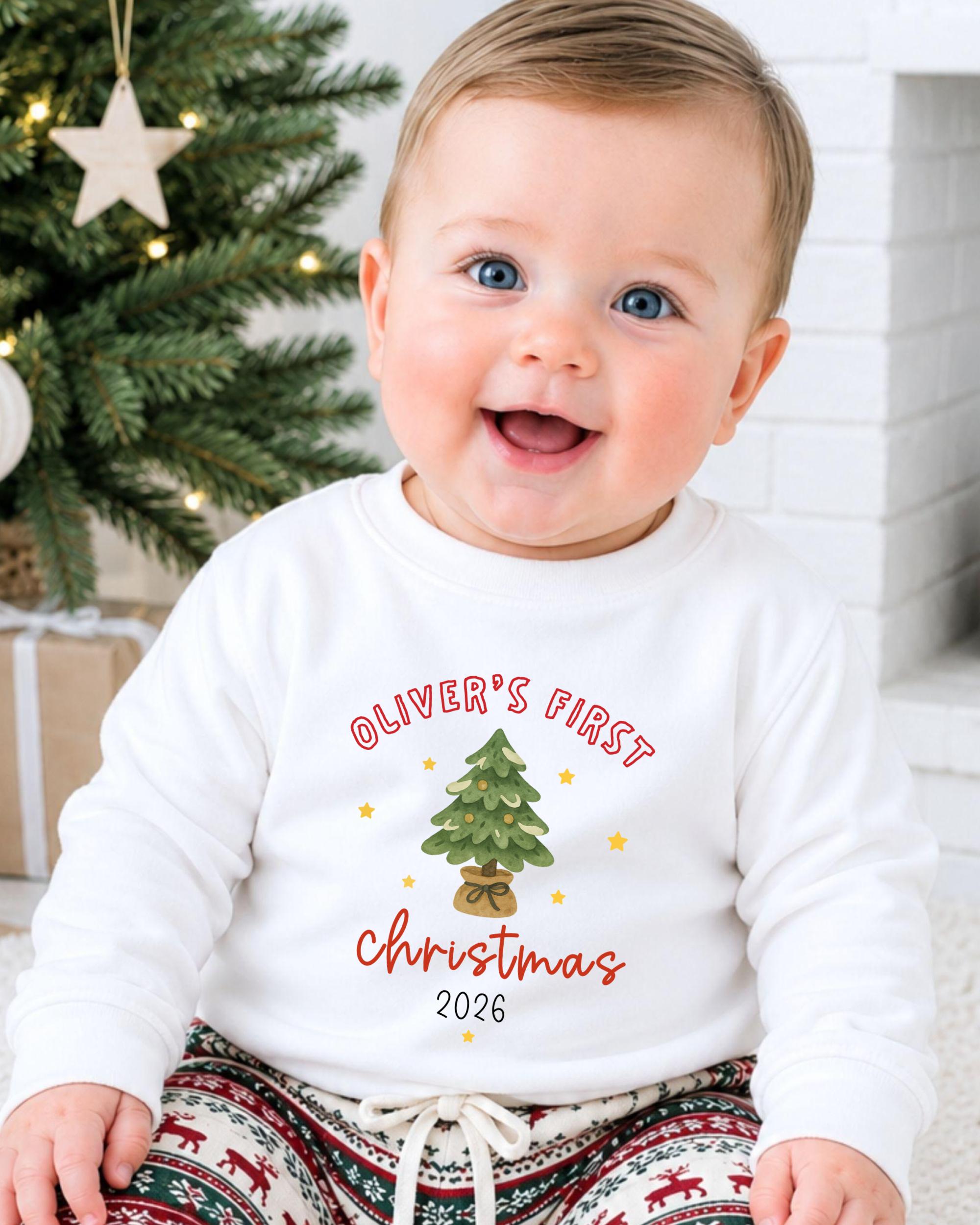 A smiling baby boy wearing a white sweatshirt with a Christmas tree design and text that says "Oliver's first Christmas 2016".