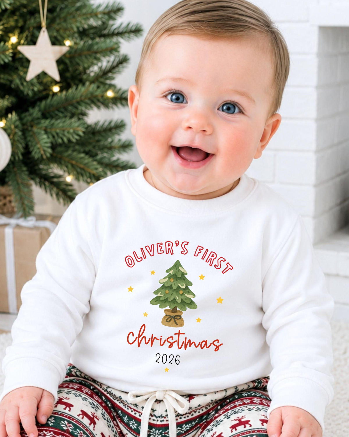 A smiling baby boy wearing a white sweatshirt with a Christmas tree design and text that says "Oliver's first Christmas 2016".