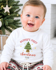 A smiling baby boy wearing a white sweatshirt with a Christmas tree design and text that says "Oliver's first Christmas 2016".