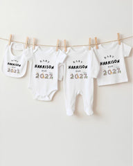 A row of white baby clothes with the text "BABY HARRISON DUE 2027" printed on them, hanging on a clothesline.