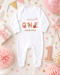 A white onesie with the text "Sweet One" and " Jennifer" printed on it, surrounded by pink and gold decorations, a pink star, and a pink balloon.