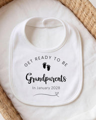 A white bib with the text "GET READY TO BE Grandparents In January 2028" printed on it, along with a small graphic of baby feet.